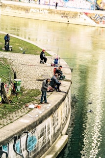 Several people are fishing along a riverbank with graffiti-covered walls. They sit and stand on a paved area, using fishing rods and assorted gear scattered around them. The water is calm, and pigeons can be seen around the fishermen.