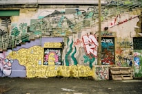 A wall covered in a variety of colorful graffiti art and street art. The wall features intricate designs, abstract patterns, and cartoonish characters. There are stairs on the left side blending into the art, and a variety of tags and text in different fonts and colors. A palette of muted and vibrant shades is visible throughout.
