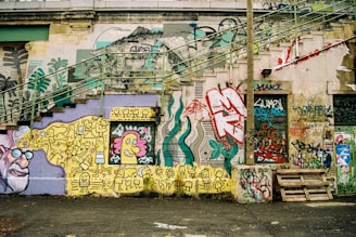 A wall covered in a variety of colorful graffiti art and street art. The wall features intricate designs, abstract patterns, and cartoonish characters. There are stairs on the left side blending into the art, and a variety of tags and text in different fonts and colors. A palette of muted and vibrant shades is visible throughout.
