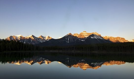 A tranquil landscape featuring snow-capped mountains bathed in golden light, reflected perfectly in the still waters of a lake. Dense forests border the lake, creating a sense of serenity and natural beauty.