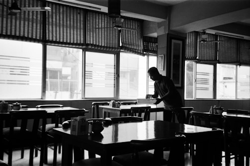A dimly lit restaurant interior has multiple wooden tables and chairs arranged in neat rows. A person is standing near a table, appearing to be cleaning or setting it. The windows have pull-down blinds that are partially closed, allowing diffused light to enter, creating a soft contrast of light and shadow throughout the room.