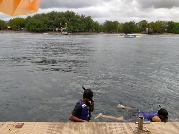Two people wearing snorkeling gear are in the water near a wooden dock. The sky is cloudy, and there is lush greenery in the background. A couple of boats are visible on the calm water, with one close to the shore.