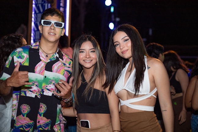 A group of three young adults at a lively event, dressed casually with vibrant attire. The man on the left wears sunglasses and a colorful shirt, while both women have long hair, one wearing a black top and the other a white wrap top. The background is dark, suggesting a night setting with bright lights.