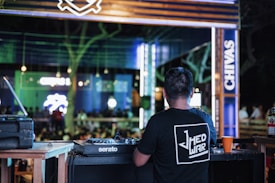 A DJ is standing at a mixing console in an outdoor venue. The setting is lively with colorful lights and a crowd in the background. Neon signs and branding are visible, creating an energetic atmosphere.