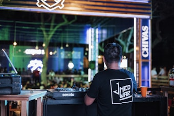 A DJ is standing at a mixing console in an outdoor venue. The setting is lively with colorful lights and a crowd in the background. Neon signs and branding are visible, creating an energetic atmosphere.