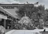 An outdoor area with various plants and garden items arranged under a large decorative structure. The setting appears to be a nursery or garden center with potted plants and other decorative items. In the background, there is a building with an American flag flying on top. A sign labeled 'hawthorne' is visible on a building structure to the left.