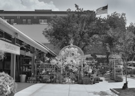 An outdoor area with various plants and garden items arranged under a large decorative structure. The setting appears to be a nursery or garden center with potted plants and other decorative items. In the background, there is a building with an American flag flying on top. A sign labeled 'hawthorne' is visible on a building structure to the left.