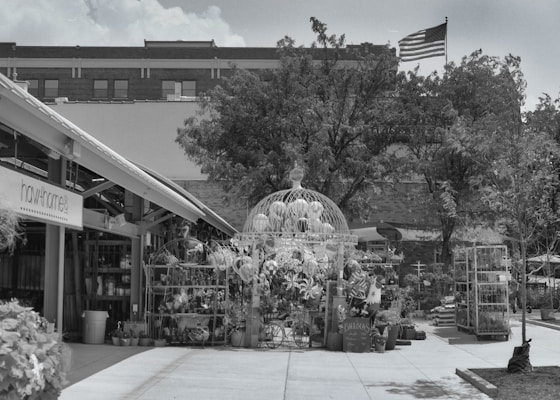 An outdoor area with various plants and garden items arranged under a large decorative structure. The setting appears to be a nursery or garden center with potted plants and other decorative items. In the background, there is a building with an American flag flying on top. A sign labeled 'hawthorne' is visible on a building structure to the left.
