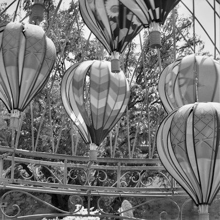 A collection of abstract wind sculptures that resemble hot air balloons with intricate patterns and designs, suspended in a framework of decorative metal arches. The background consists of leafy trees, adding an organic contrast to the geometric shapes of the balloons.
