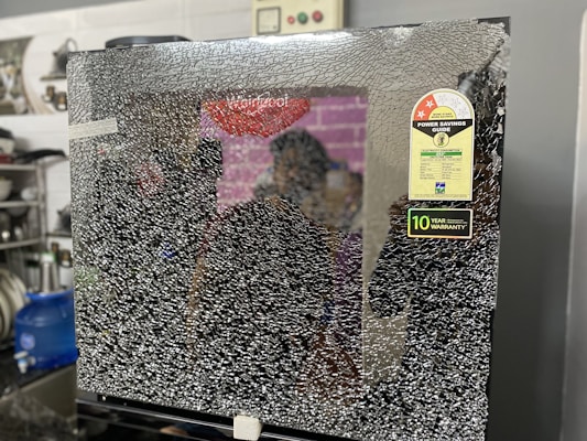 A shattered glass panel with a sticker labeled 'Power Savings Guide' and a note indicating a 10-year warranty. The background includes a kitchen setting with a water dispenser, dishes, and some utensils.