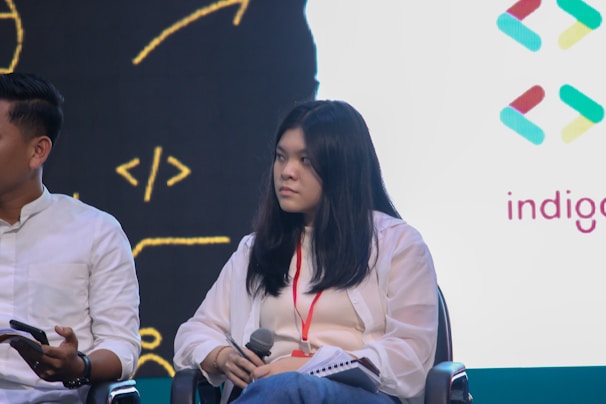 A woman with long dark hair holds a microphone while sitting on a chair, wearing a white blouse and a red lanyard. She looks to the side attentively, and there is a logo with geometric shapes and the word 'indigo' visible on a white background. To her left, a person wearing a white shirt holds a smartphone.