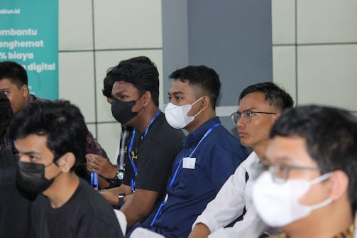 A group of people are seated in a row, wearing masks due to health precautions. Some are dressed in casual shirts, and one person is wearing glasses. They appear to be attentively listening, suggesting a seminar, workshop, or meeting setting.
