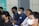 A group of people are seated in a row, wearing masks due to health precautions. Some are dressed in casual shirts, and one person is wearing glasses. They appear to be attentively listening, suggesting a seminar, workshop, or meeting setting.