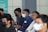 A group of people are seated in a row, wearing masks due to health precautions. Some are dressed in casual shirts, and one person is wearing glasses. They appear to be attentively listening, suggesting a seminar, workshop, or meeting setting.