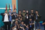 A group of young people, mostly men, gather together in a room posed for a friendly group photo. Many are wearing casual clothing and identification badges around their necks. Some individuals have expressions of happiness, giving thumbs-up gestures. Behind them are multiple flags and a blank screen, suggesting a possible event or conference setting.