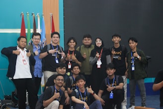A group of young people, mostly men, gather together in a room posed for a friendly group photo. Many are wearing casual clothing and identification badges around their necks. Some individuals have expressions of happiness, giving thumbs-up gestures. Behind them are multiple flags and a blank screen, suggesting a possible event or conference setting.