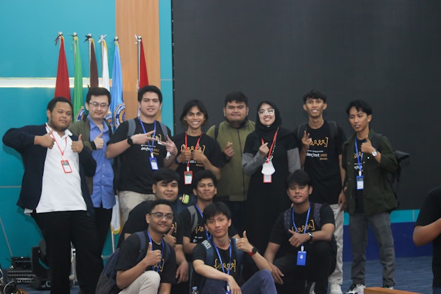 A group of young people, mostly men, gather together in a room posed for a friendly group photo. Many are wearing casual clothing and identification badges around their necks. Some individuals have expressions of happiness, giving thumbs-up gestures. Behind them are multiple flags and a blank screen, suggesting a possible event or conference setting.