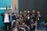 A group of young people, mostly men, gather together in a room posed for a friendly group photo. Many are wearing casual clothing and identification badges around their necks. Some individuals have expressions of happiness, giving thumbs-up gestures. Behind them are multiple flags and a blank screen, suggesting a possible event or conference setting.