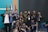 A group of young people, mostly men, gather together in a room posed for a friendly group photo. Many are wearing casual clothing and identification badges around their necks. Some individuals have expressions of happiness, giving thumbs-up gestures. Behind them are multiple flags and a blank screen, suggesting a possible event or conference setting.