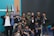 A group of young people, mostly men, gather together in a room posed for a friendly group photo. Many are wearing casual clothing and identification badges around their necks. Some individuals have expressions of happiness, giving thumbs-up gestures. Behind them are multiple flags and a blank screen, suggesting a possible event or conference setting.