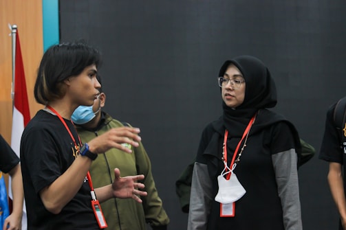 Two people are engaged in a conversation. One person, wearing a black shirt and a lanyard, is gesturing with their hands, while the other person, in a black hijab and glasses, listens attentively. A red and white flag is partially visible in the background.