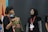 Two people are engaged in a conversation. One person, wearing a black shirt and a lanyard, is gesturing with their hands, while the other person, in a black hijab and glasses, listens attentively. A red and white flag is partially visible in the background.