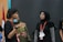 Two people are engaged in a conversation. One person, wearing a black shirt and a lanyard, is gesturing with their hands, while the other person, in a black hijab and glasses, listens attentively. A red and white flag is partially visible in the background.