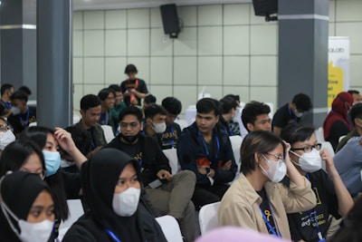 A group of seated individuals, mostly wearing casual attire with matching lanyards, are gathered in what appears to be a conference or seminar setting. Many are wearing face masks and seem engaged or attentive, possibly listening to a speaker outside the frame. The setting is indoors with neutral-colored walls and several attendees are visible in a medium-sized room.