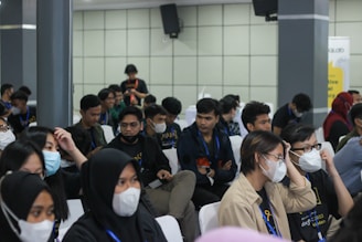 A group of seated individuals, mostly wearing casual attire with matching lanyards, are gathered in what appears to be a conference or seminar setting. Many are wearing face masks and seem engaged or attentive, possibly listening to a speaker outside the frame. The setting is indoors with neutral-colored walls and several attendees are visible in a medium-sized room.