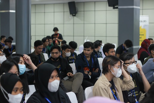 A group of seated individuals, mostly wearing casual attire with matching lanyards, are gathered in what appears to be a conference or seminar setting. Many are wearing face masks and seem engaged or attentive, possibly listening to a speaker outside the frame. The setting is indoors with neutral-colored walls and several attendees are visible in a medium-sized room.