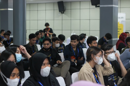 A group of seated individuals, mostly wearing casual attire with matching lanyards, are gathered in what appears to be a conference or seminar setting. Many are wearing face masks and seem engaged or attentive, possibly listening to a speaker outside the frame. The setting is indoors with neutral-colored walls and several attendees are visible in a medium-sized room.