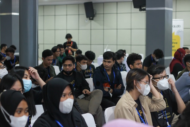 A group of seated individuals, mostly wearing casual attire with matching lanyards, are gathered in what appears to be a conference or seminar setting. Many are wearing face masks and seem engaged or attentive, possibly listening to a speaker outside the frame. The setting is indoors with neutral-colored walls and several attendees are visible in a medium-sized room.