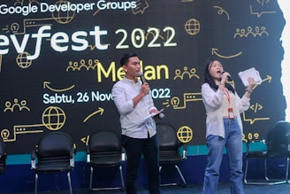 Two presenters stand on stage holding microphones, with an audience behind them in chairs. The backdrop features text and graphics related to a developer conference called 'Devfest 2022' along with date and location details.