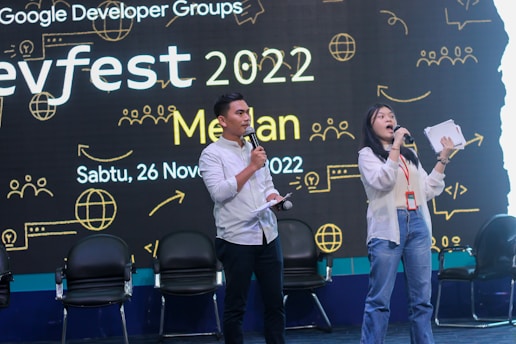 Two presenters stand on stage holding microphones, with an audience behind them in chairs. The backdrop features text and graphics related to a developer conference called 'Devfest 2022' along with date and location details.