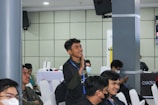 A group of young individuals is seated in a conference or seminar room. One person is standing and appears to be actively engaging or making a gesture, possibly speaking or asking a question. Others are sitting on white chairs, some wearing face masks, indicating attentiveness or interest. The background features a speaker mounted on the wall and a table with promotional banners.