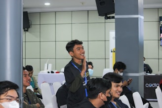A group of young individuals is seated in a conference or seminar room. One person is standing and appears to be actively engaging or making a gesture, possibly speaking or asking a question. Others are sitting on white chairs, some wearing face masks, indicating attentiveness or interest. The background features a speaker mounted on the wall and a table with promotional banners.