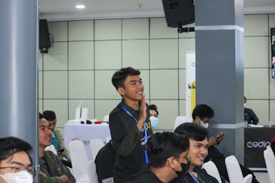 A group of young individuals is seated in a conference or seminar room. One person is standing and appears to be actively engaging or making a gesture, possibly speaking or asking a question. Others are sitting on white chairs, some wearing face masks, indicating attentiveness or interest. The background features a speaker mounted on the wall and a table with promotional banners.