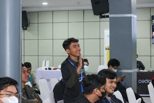 A group of young individuals is seated in a conference or seminar room. One person is standing and appears to be actively engaging or making a gesture, possibly speaking or asking a question. Others are sitting on white chairs, some wearing face masks, indicating attentiveness or interest. The background features a speaker mounted on the wall and a table with promotional banners.