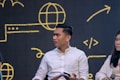 A man in a white shirt is sitting with a digital gadget in his hand, while a woman next to him holds a microphone. They are in front of a dark backdrop with yellow digital symbols like a globe and code arrows.