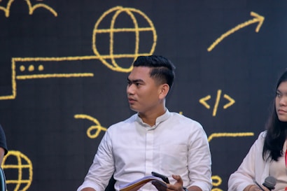 A man in a white shirt is sitting with a digital gadget in his hand, while a woman next to him holds a microphone. They are in front of a dark backdrop with yellow digital symbols like a globe and code arrows.