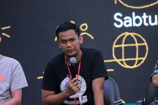 A person holding a microphone appears to be speaking or presenting. They are seated, wearing a black t-shirt with a design on it. In the background, there is a dark backdrop with abstract yellow line art and text including the word 'Sabtu'.