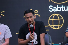A person holding a microphone appears to be speaking or presenting. They are seated, wearing a black t-shirt with a design on it. In the background, there is a dark backdrop with abstract yellow line art and text including the word 'Sabtu'.