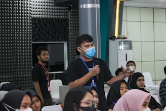 Several people are seated in a room listening attentively. One person stands holding a microphone, appearing to ask a question or make a comment. Most individuals are wearing face masks, indicating a likely indoor event during a health-conscious period. The room has a modern design with paneling on the walls and an air conditioning unit visible.