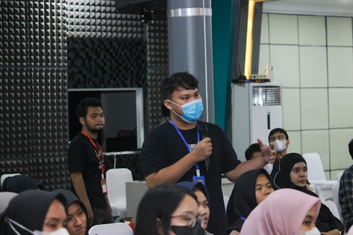Several people are seated in a room listening attentively. One person stands holding a microphone, appearing to ask a question or make a comment. Most individuals are wearing face masks, indicating a likely indoor event during a health-conscious period. The room has a modern design with paneling on the walls and an air conditioning unit visible.