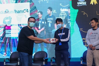 A group of people on a stage, with one person handing an item to another. They are wearing casual clothing and face masks. The background features a large screen displaying text and graphics.