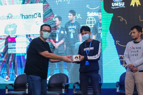 A group of people on a stage, with one person handing an item to another. They are wearing casual clothing and face masks. The background features a large screen displaying text and graphics.