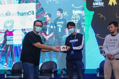A group of people on a stage, with one person handing an item to another. They are wearing casual clothing and face masks. The background features a large screen displaying text and graphics.