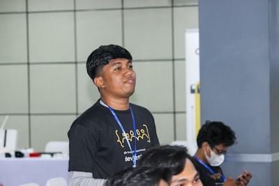 A young person with short dark hair is wearing a black t-shirt with yellow print and a blue lanyard. They appear to be standing indoors against a tiled wall background. In the foreground, part of another person is visible, and in the background, another individual is seated and looking down at an object, possibly a smartphone. The setting appears to be a casual or professional gathering or event.