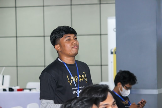 A young person with short dark hair is wearing a black t-shirt with yellow print and a blue lanyard. They appear to be standing indoors against a tiled wall background. In the foreground, part of another person is visible, and in the background, another individual is seated and looking down at an object, possibly a smartphone. The setting appears to be a casual or professional gathering or event.