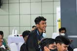 Several young individuals are sitting in a classroom or conference setting. One person is standing and smiling, while others are seated, some wearing masks. The atmosphere appears to be informal and engaging.